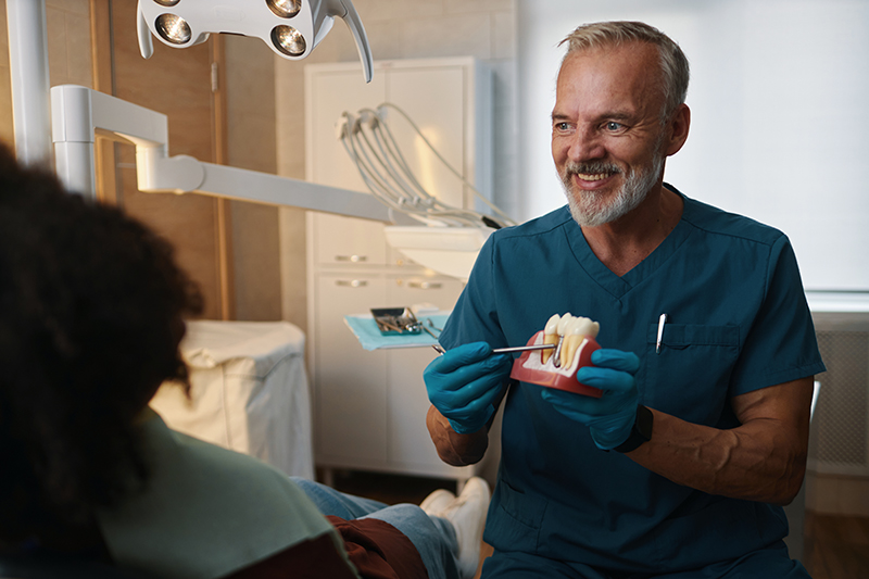 Smiling dentist holding dental model while explaining procedure to patient in clinic setting with medical equipment and dental chair visible in background